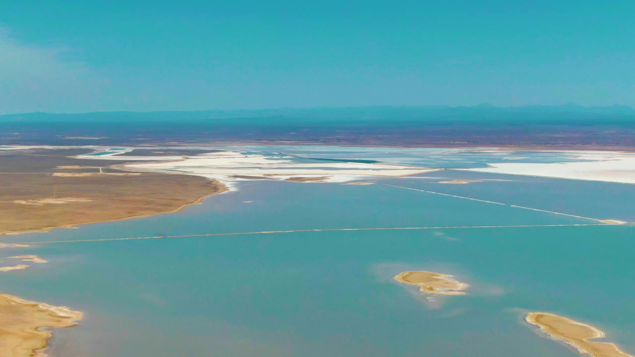 One second into the clip, the aerial view in Guerrero Negro holds on blue-green water, a bright white salt-flat edge, tan shoreline, distant low mountains, and no visible people.