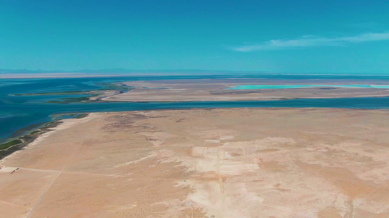 Opening aerial frame over Guerrero Negro, Baja California Sur, with pale sandy desert in front, a calm blue water channel beyond it, faint mountains on the horizon, and no visible people.