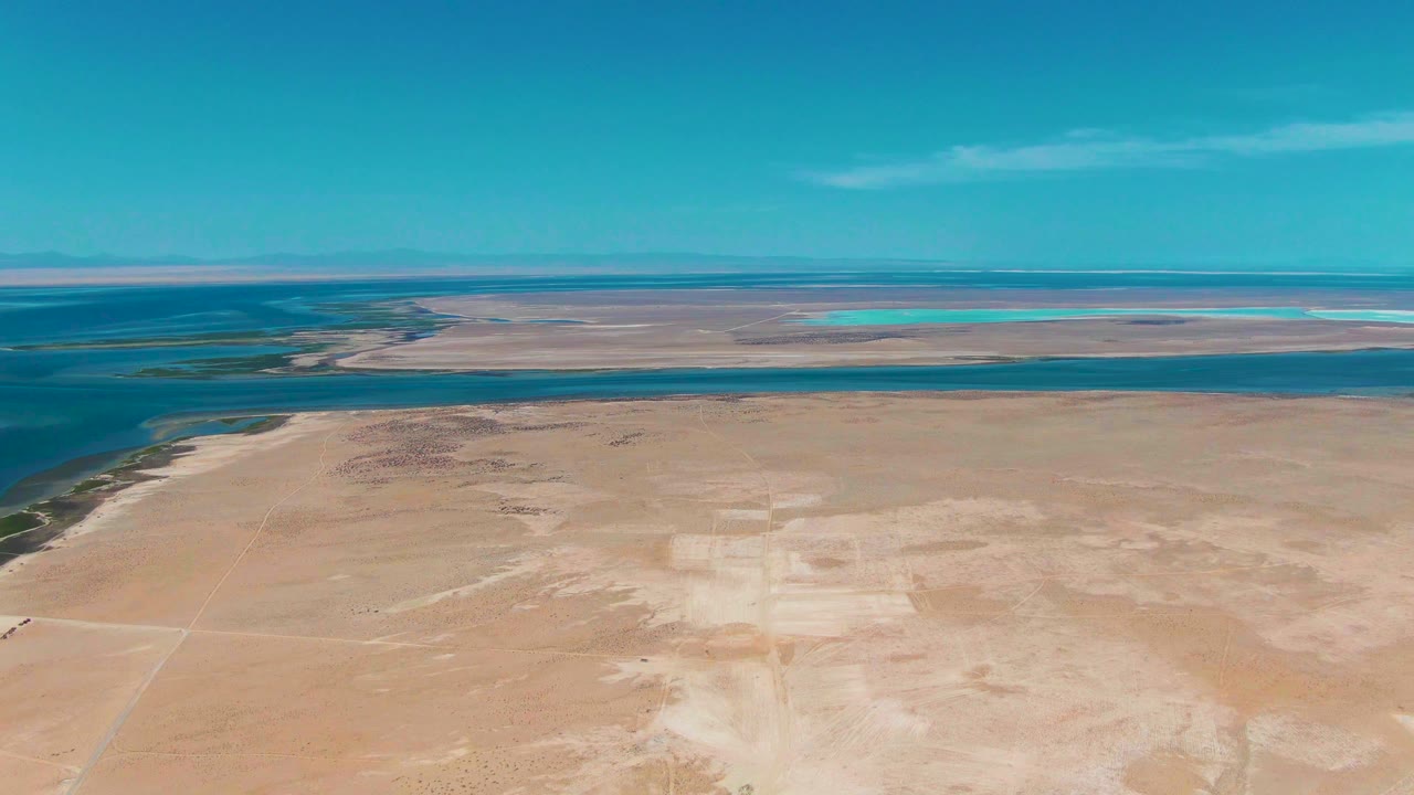 Second frame over Guerrero Negro shows the sandy foreground slightly advanced in the composition, with calm water bands, distant turquoise pools, a clear sky, and no visible people.