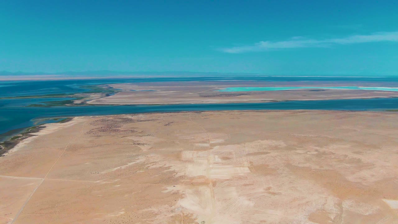 Third frame in Guerrero Negro holds the desert foreground, dark blue midground water channel, pale distant flats, turquoise water patches, clear blue sky, and no visible people.