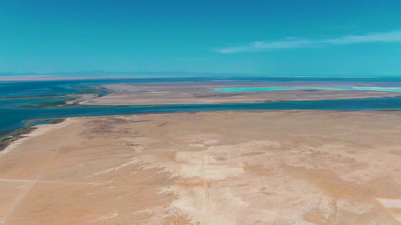 Fourth frame over Guerrero Negro shows more of the left shoreline curve, sandy desert across the lower frame, calm blue water in the middle distance, hazy mountains, and no visible people.