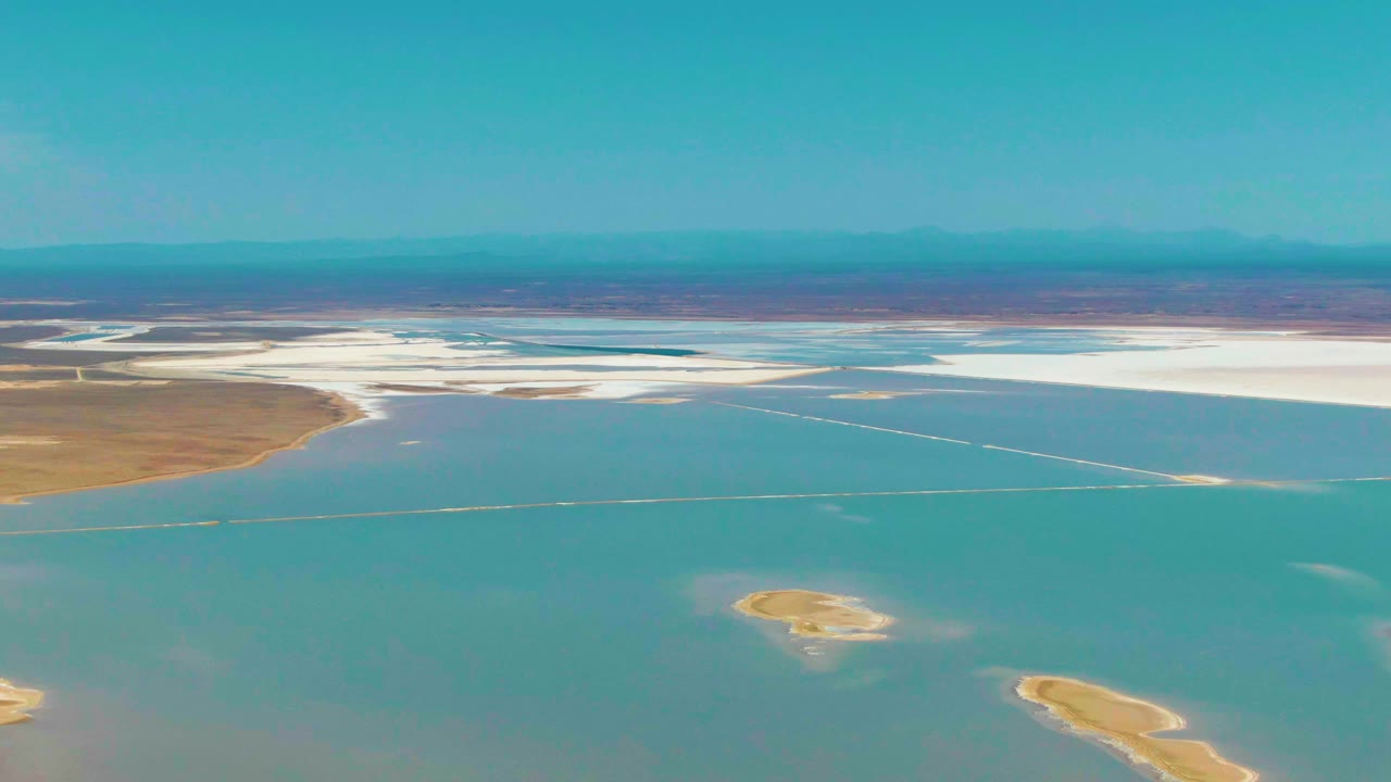 Aerial frame in Guerrero Negro, Baja California Sur showing turquoise shallow water, small tan islands closer to the bottom edge, distant white salt flats, and no visible people.