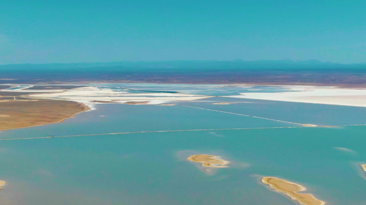 Aerial view over Guerrero Negro, Baja California Sur with a broad band of blue water, white salt flats across the background, brown land at left, small islands below, and no visible people.