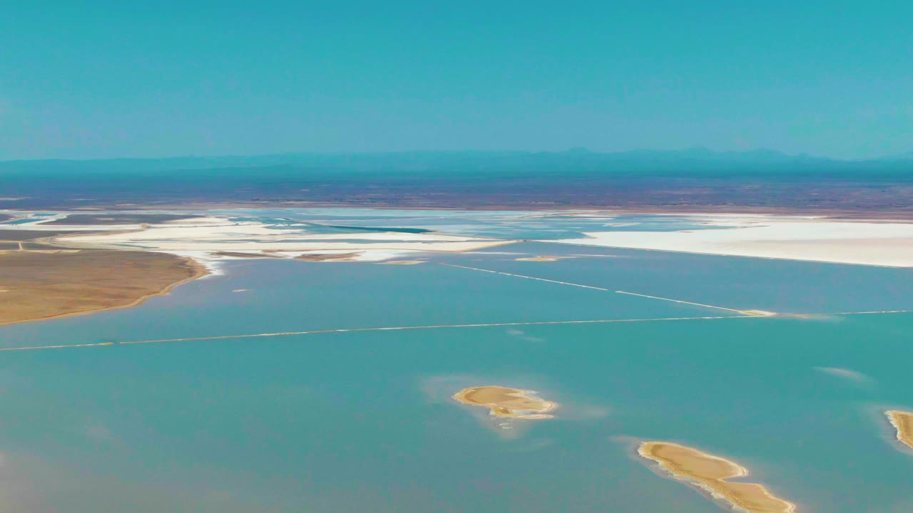 Mid-clip aerial frame from Guerrero Negro, Baja California Sur showing blue water, a small tan island near the lower center, white salt flats beyond, and no visible people.