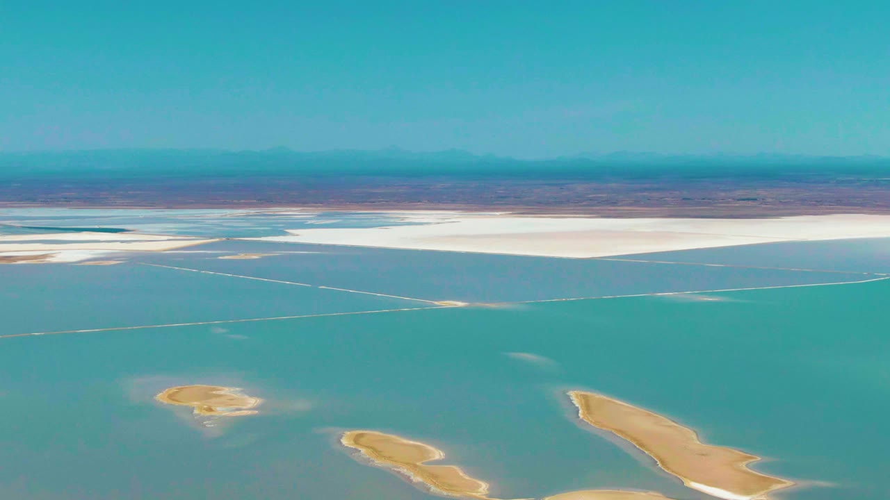 Second aerial frame over Guerrero Negro shows shallow turquoise water crossing the midground, white salt flats stretching along the far shore, foreground sandbars, distant mountains, and no visible people.