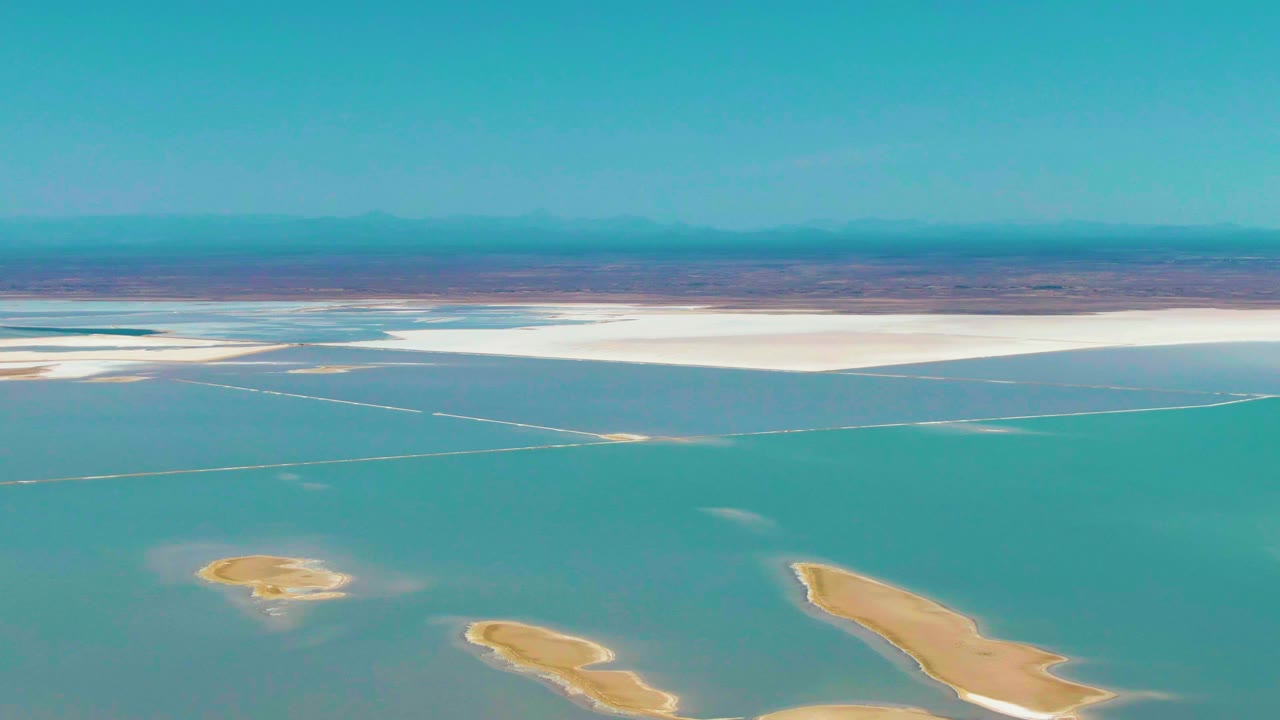Fourth frame over Guerrero Negro shows turquoise water filling most of the view, small sandy islands near the lower edge, a white salt-flat band at the far side, hazy mountains, and no visible people.