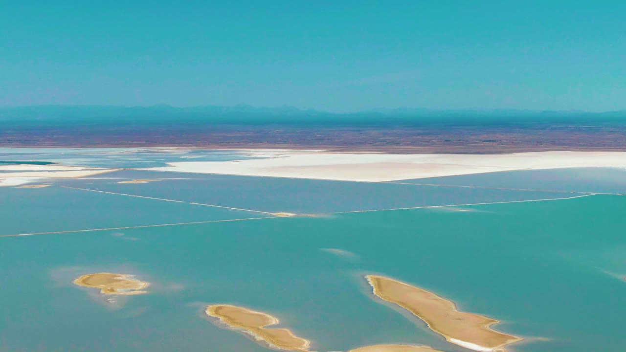 Opening aerial frame over Guerrero Negro with teal shallow water, tan exposed lake bed islands, a white salt flat band, distant mountains, and no visible people.