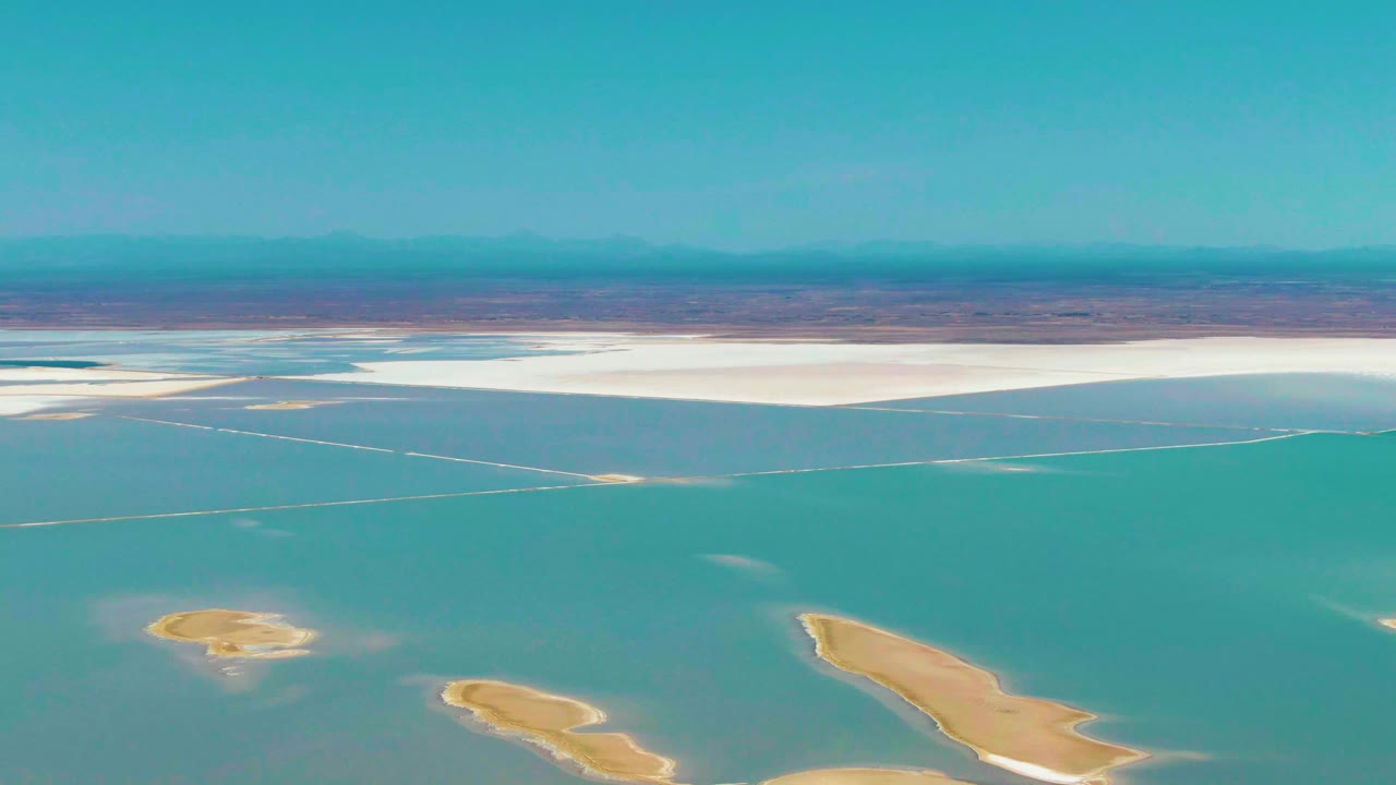 Aerial view in Guerrero Negro showing turquoise shallow water around sandy landmasses, a bright salt flat across the midground, hazy mountains, and no visible people.