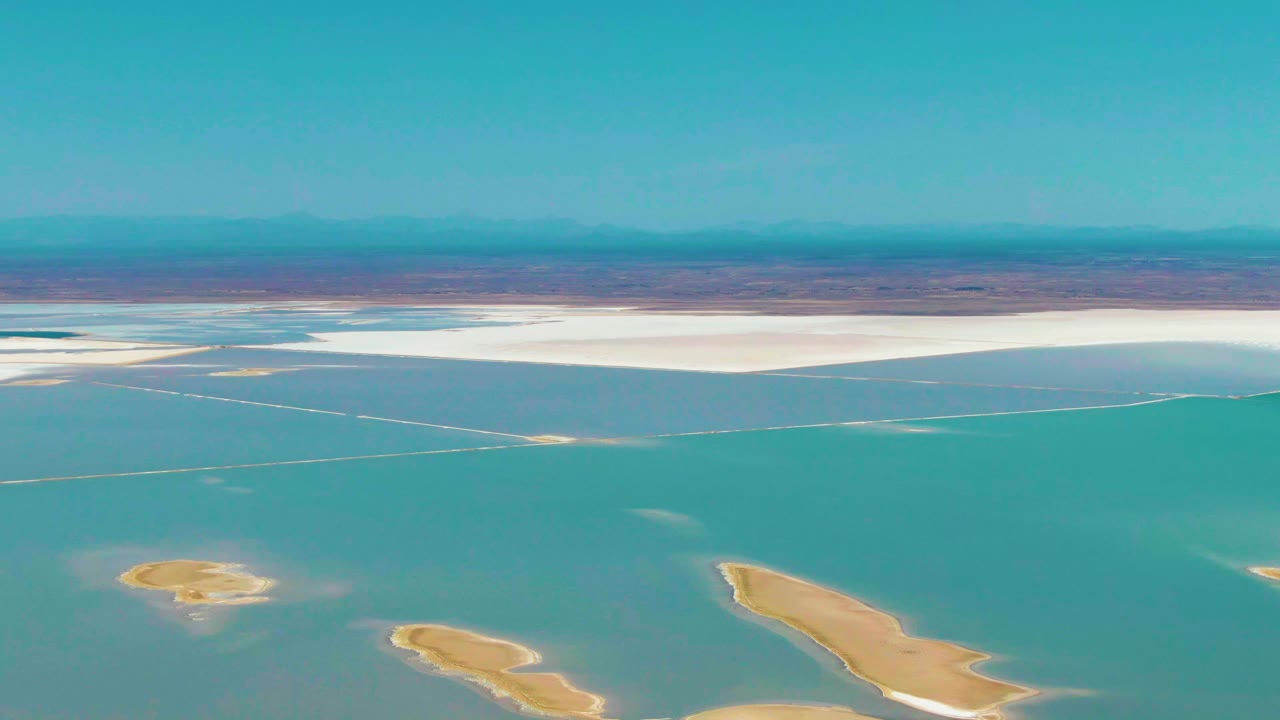 Guerrero Negro aerial frame with teal water in the foreground, two small tan islands near the lower edge, a white salt flat in the distance, mountains, and no visible people.