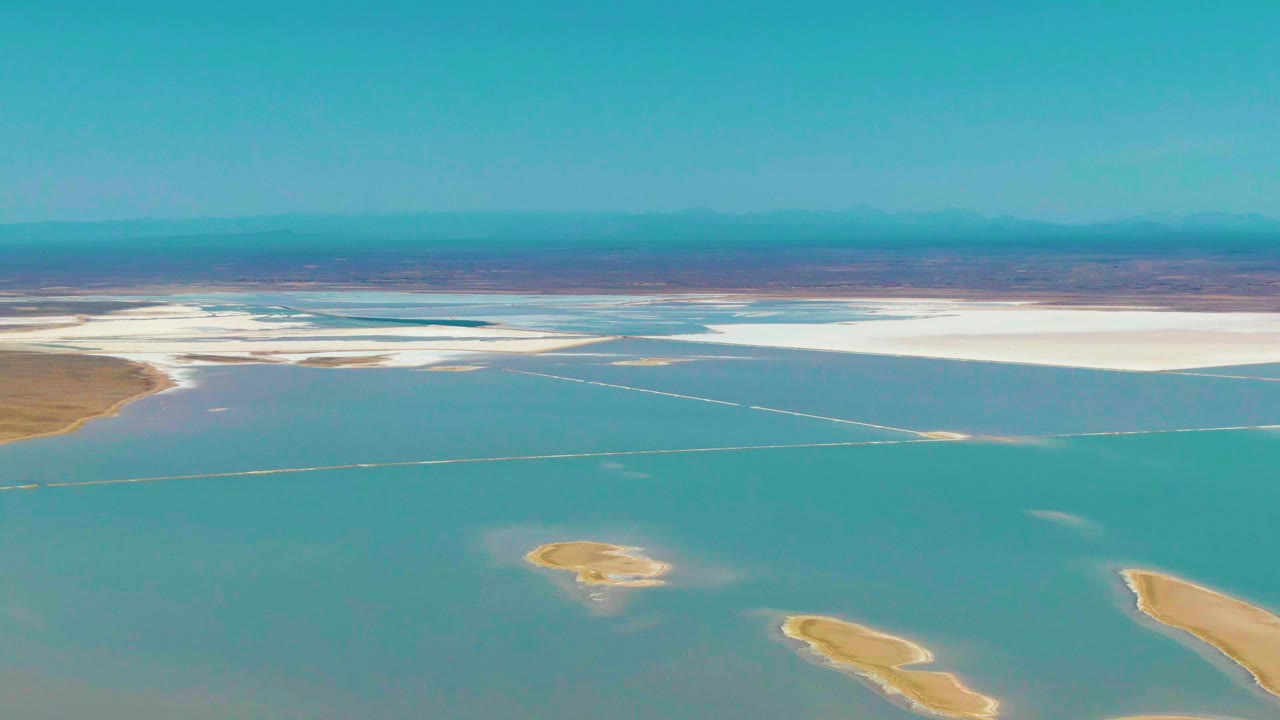 At 0:00, an aerial view over Guerrero Negro shows turquoise shallow water, white salt flats across the far shore, tan sandbars below, hazy mountains on the horizon, and no visible people.