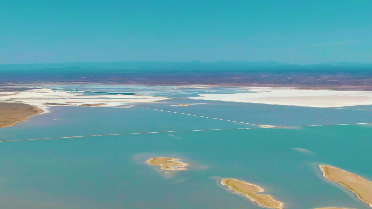At 0:01, the Guerrero Negro aerial frame holds on turquoise lagoon water with a bright white salt-flat band, small tan islands near the lower edge, distant mountains, and no visible people.