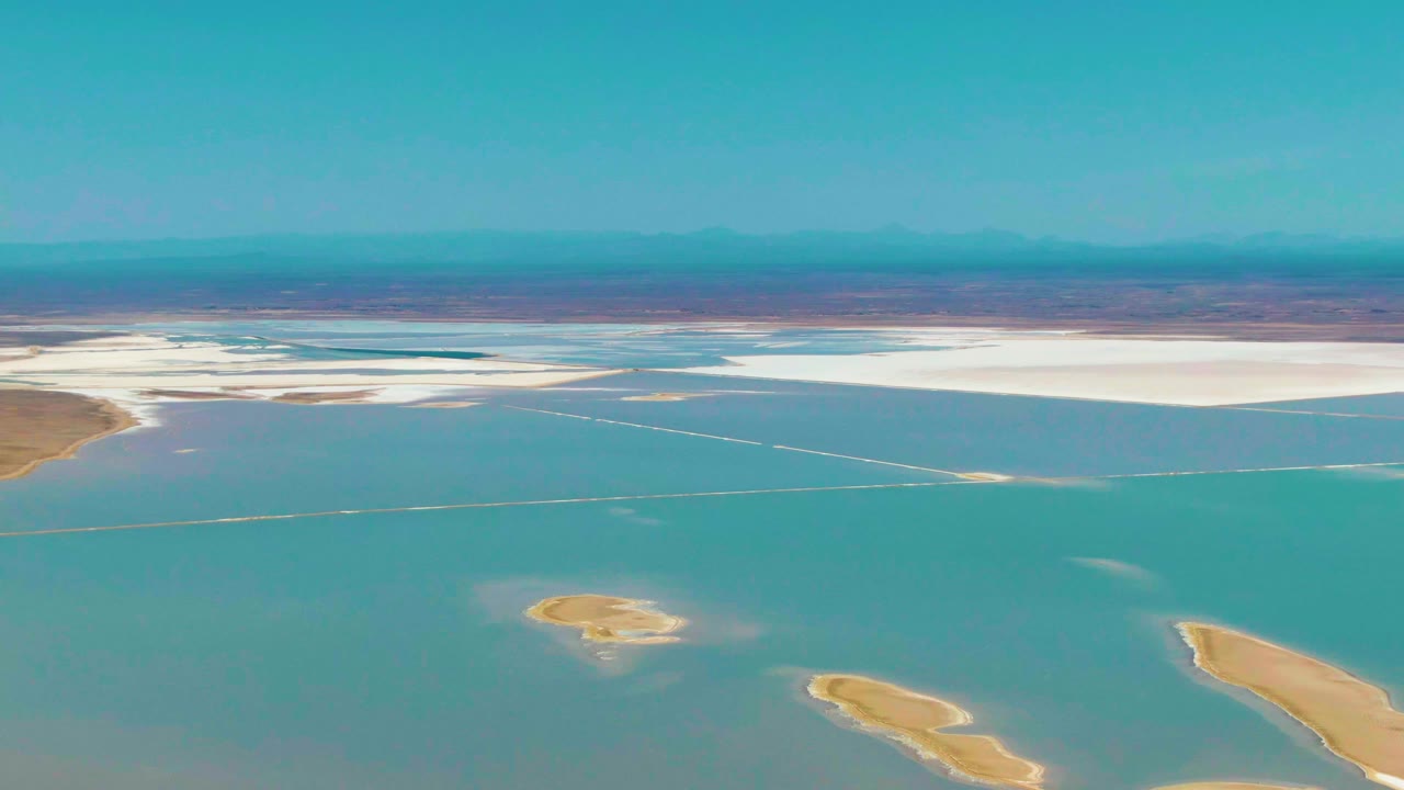 At 0:02, the aerial view in Guerrero Negro shows broad turquoise water below a pale salt-flat shelf, tan landforms entering from the lower right, hazy mountains, and no visible people.