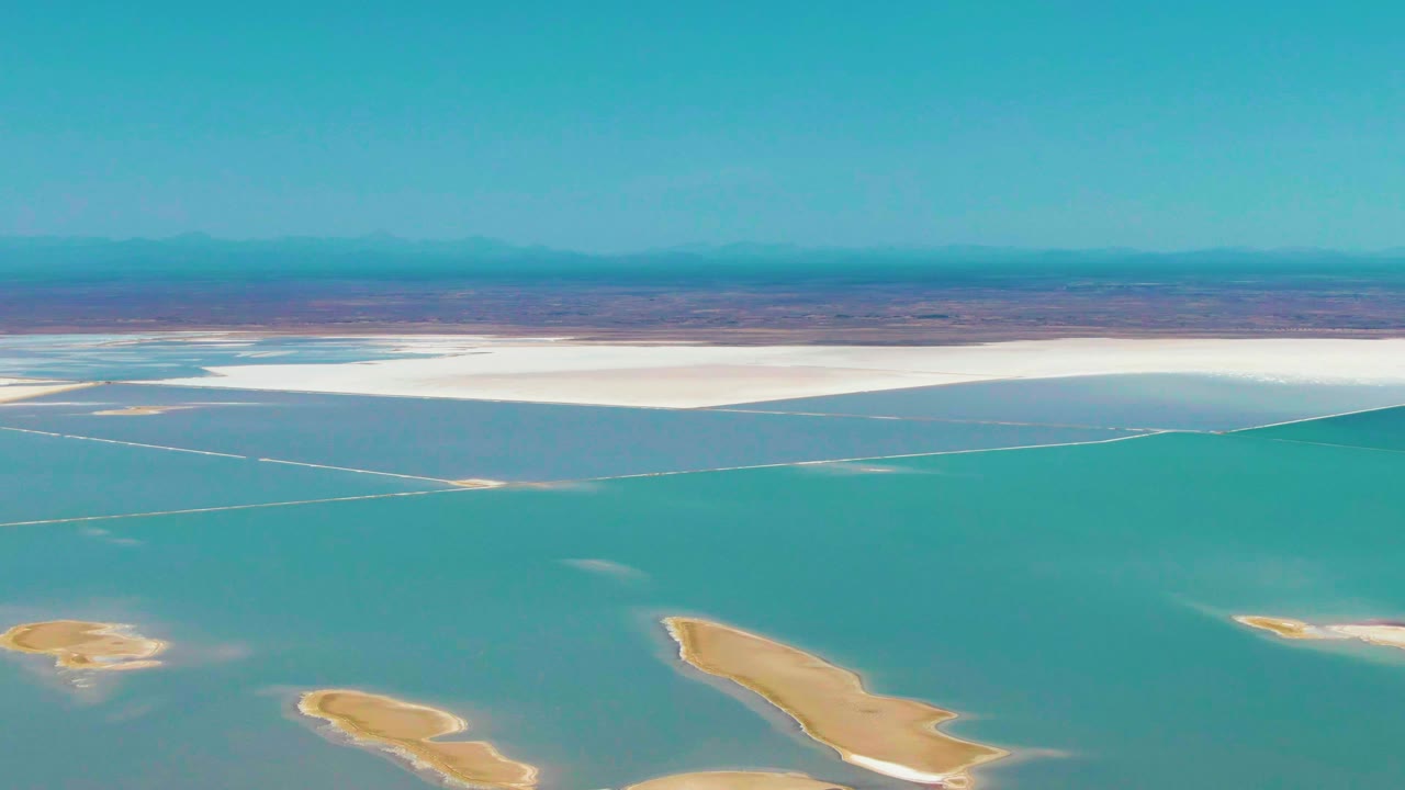 Aerial frame from Guerrero Negro, Baja California Sur showing turquoise water in the foreground, tan sand islands near the bottom edge, white salt flats across the far shore, distant dry land, and no visible people.