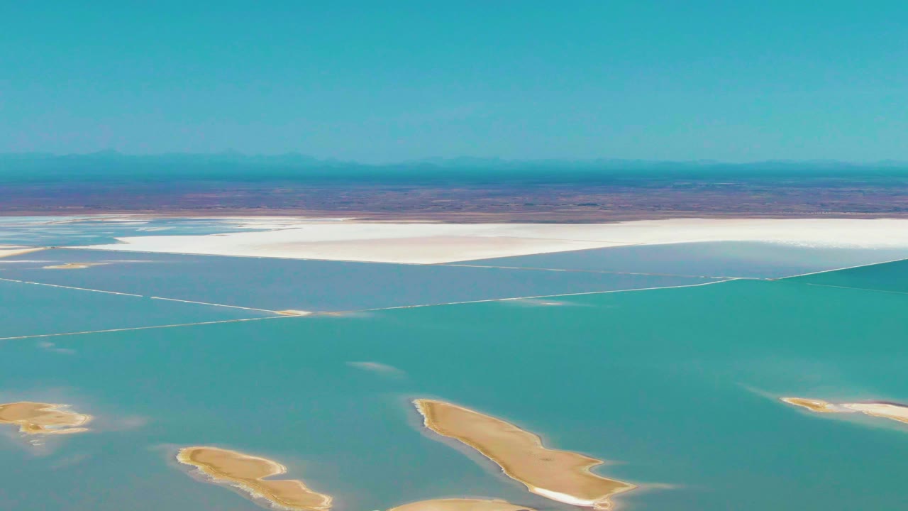 Second frame from Guerrero Negro, Baja California Sur with a broad turquoise water surface, small pale islands lower in frame, a bright white salt flat band along the shore, reddish desert beyond, and no visible people.