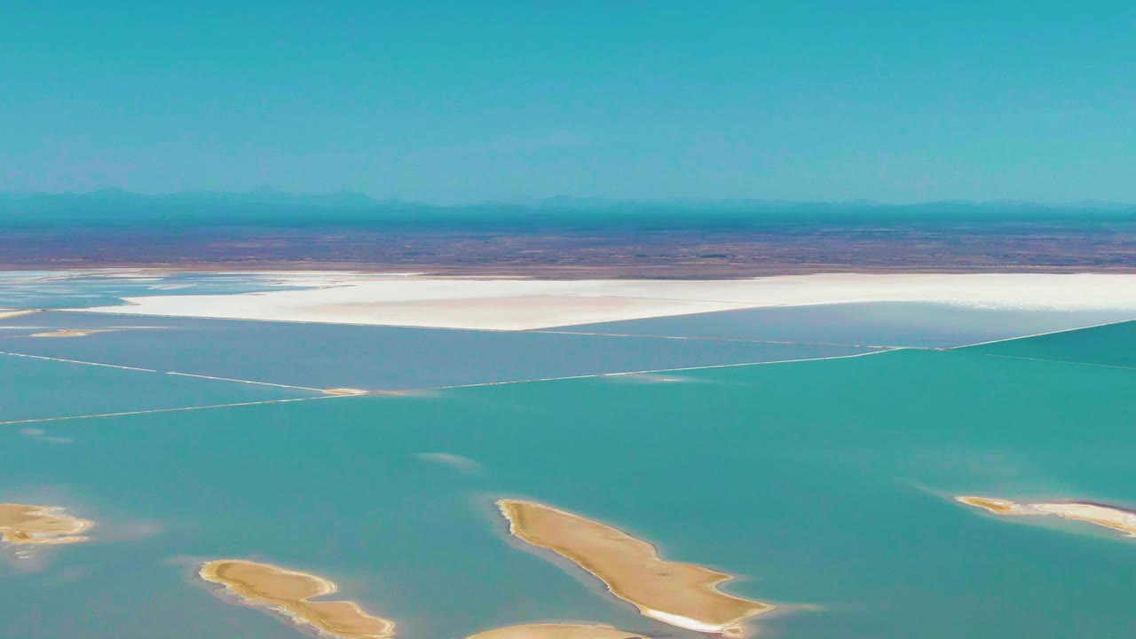 Mid-clip aerial frame in Guerrero Negro, Baja California Sur showing turquoise lagoon water, two exposed tan islands near the lower frame, a continuous white salt flat strip, distant desert terrain, and no visible people.