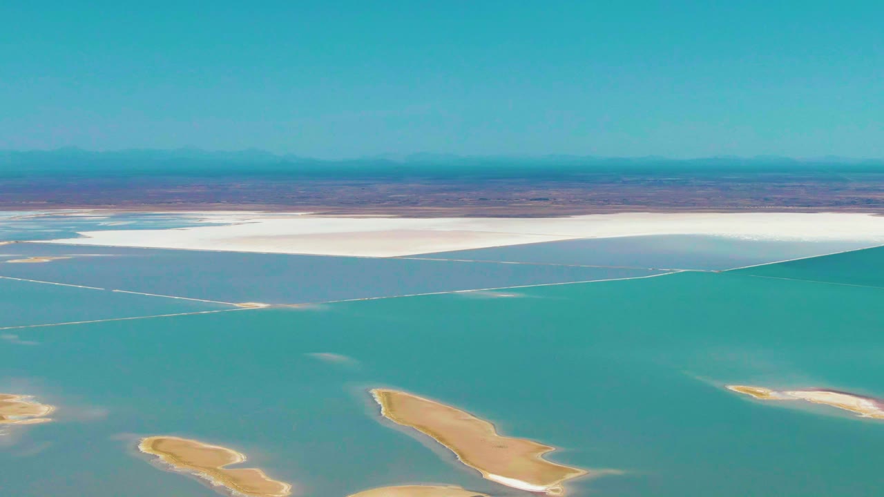 Later aerial frame from Guerrero Negro, Baja California Sur with turquoise water covering most of the view, tan sand bars along the lower edge, bright white salt flats at the far shore, hazy mountains, and no visible people.