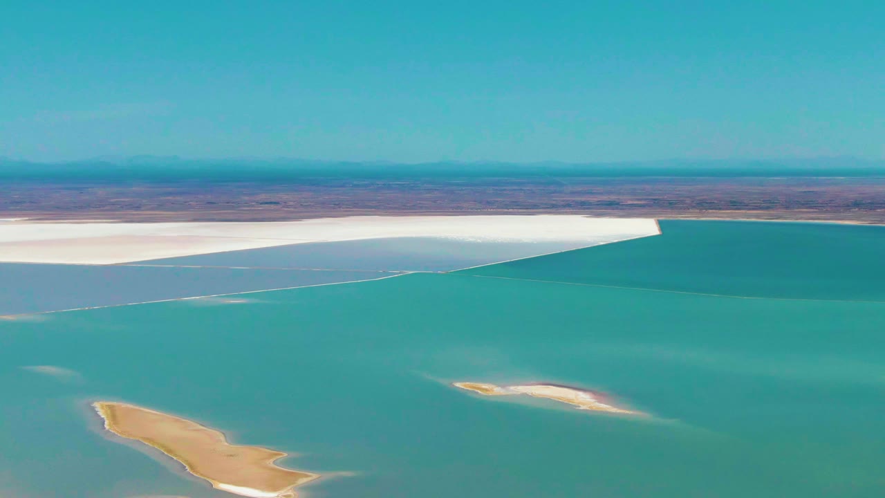 Turquoise water curves around small sand islands below a white salt flat.
