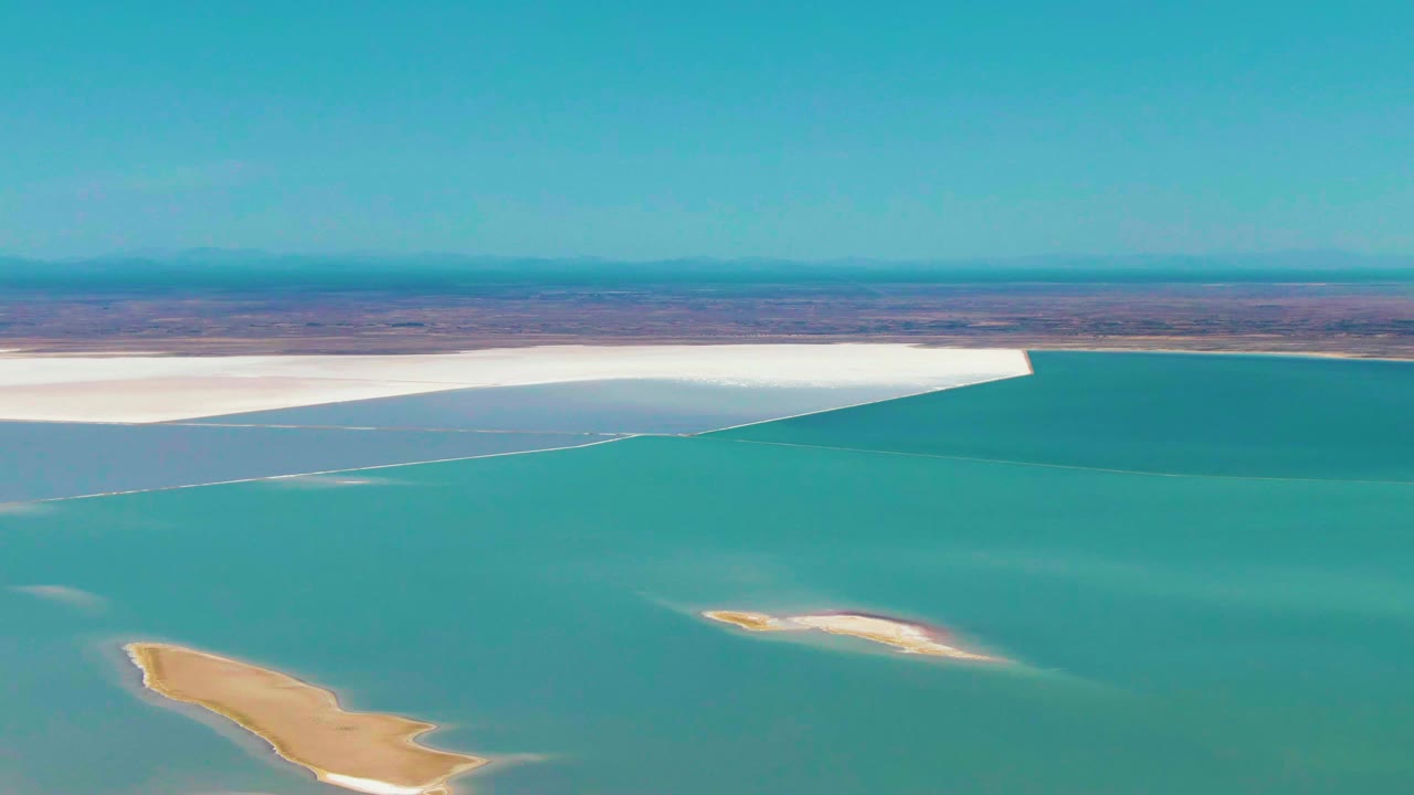 Wide aerial view in Guerrero Negro, Baja California Sur, with a level desert horizon, white salt flat, turquoise water, tan sand islands, and no visible people.