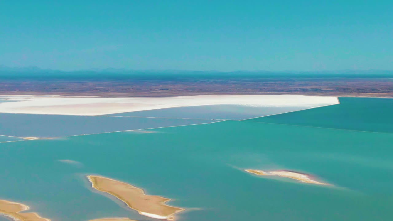 Opening aerial frame over Guerrero Negro turquoise lagoon water with sandy islets in the lower frame, a white salt flat across the shore, distant brown land, blue sky, and no visible people.