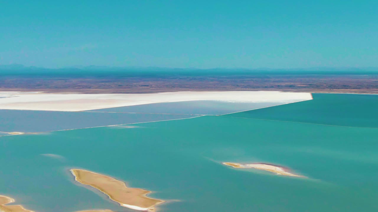 Two seconds into the clip, Guerrero Negro lagoon water fills most of the frame, with sandy islets near the bottom, a bright white salt flat along the far side, distant brown terrain, and no visible people.