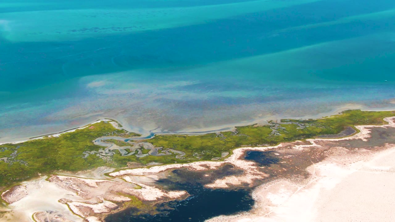 High aerial view of blue ocean bands and winding wetland channels beside pale sand.