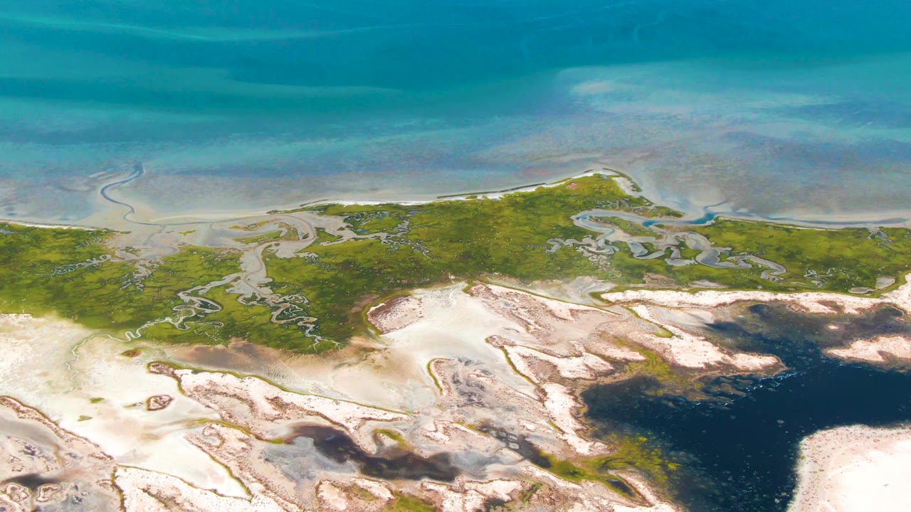 Aerial frame over Guerrero Negro, Baja California Sur, with clear shallow water above the marsh edge, branching tidal channels, pale sand flats, and no visible people.