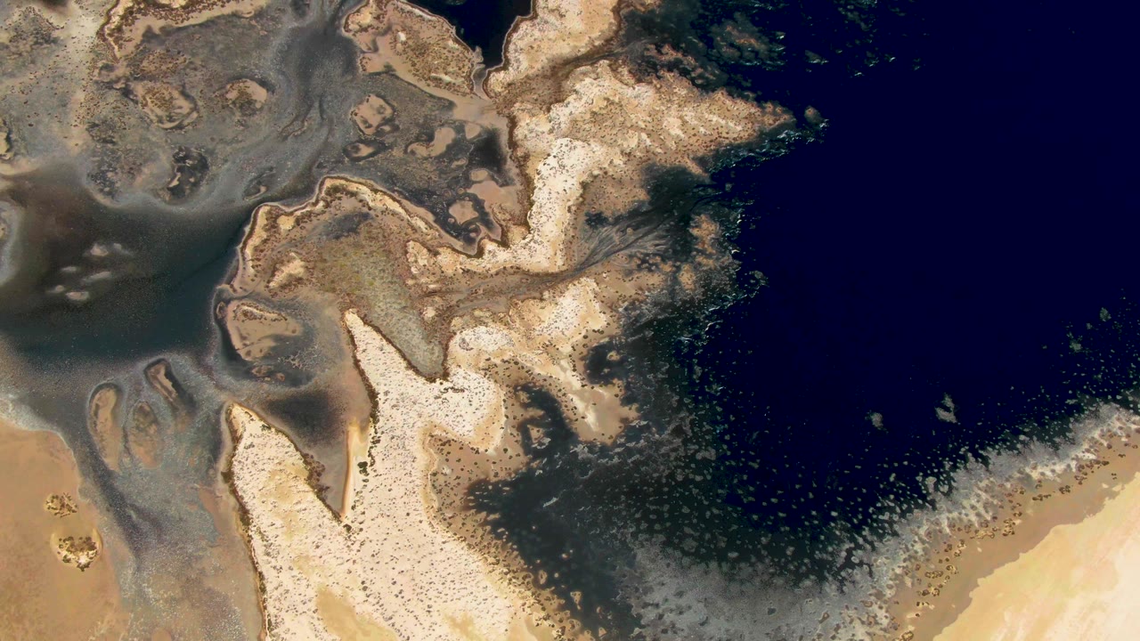 Aerial frame from Guerrero Negro, Baja California Sur, with deep blue water on the right, light sandy landforms below, and cloudy sediment spreading along the shore, with no visible people.