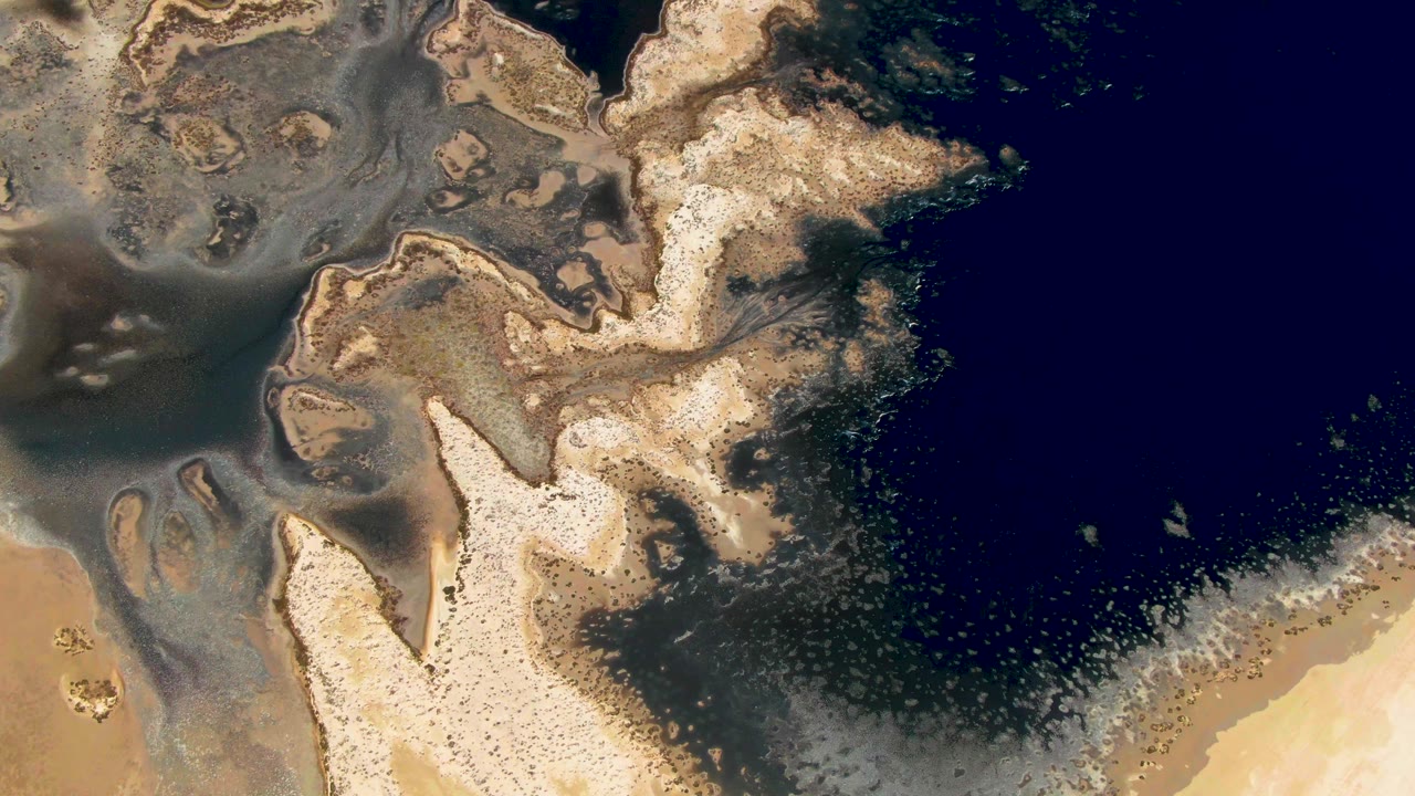 Overhead coastal view in Guerrero Negro, Baja California Sur, showing pale sandbars, brown-gray shallows, and a large dark blue water area, with no visible people.