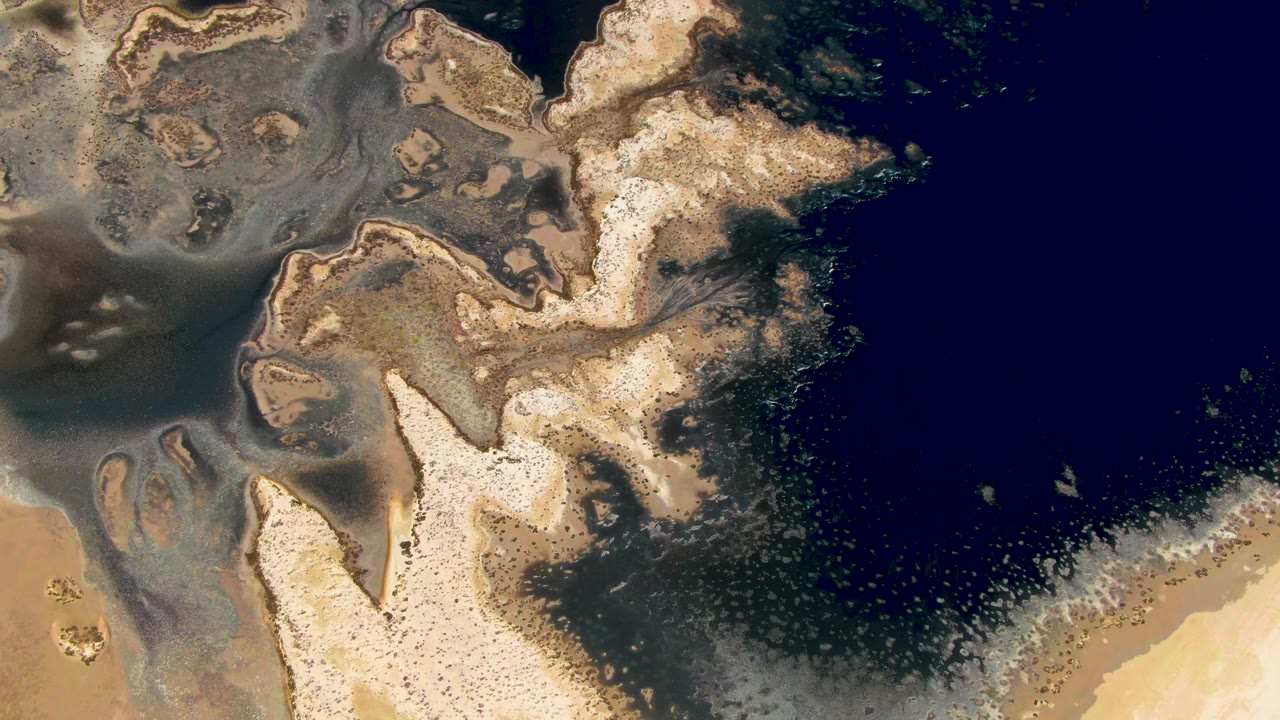Overhead frame in Guerrero Negro, Baja California Sur, with a pale sandy point, mottled brown sediment, gray-green shallow water, and dark blue water to the right, with no visible people.