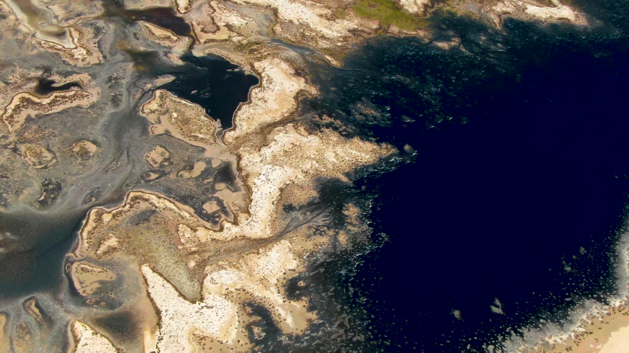 Overhead Guerrero Negro coastline frame with a broad dark water field on the right, irregular pale sandbars through the center, and no visible people.