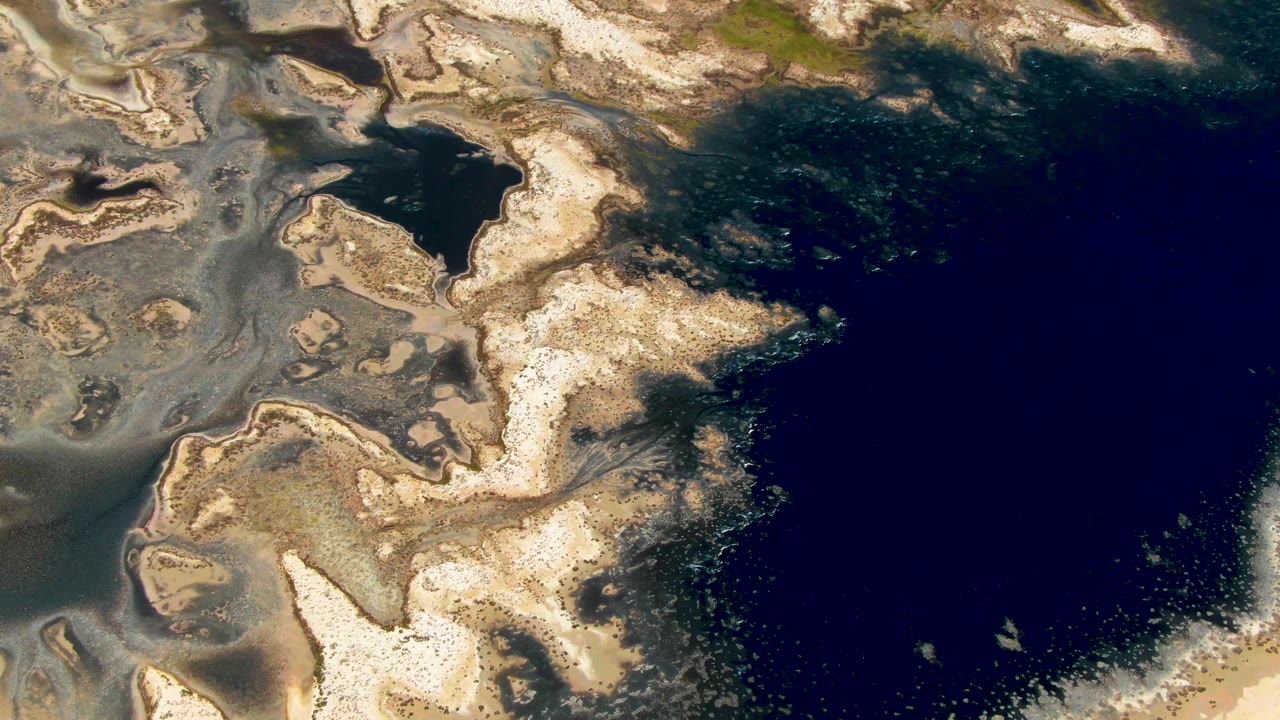 Overhead view of Guerrero Negro tidal water with more foamy speckles along the lower right edge, layered beige sediment across the center, and no visible people.