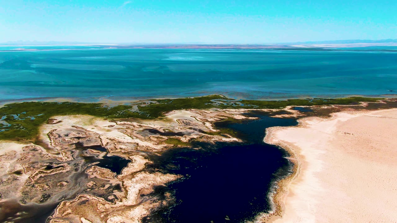 Opening aerial frame over Guerrero Negro with a dark lagoon below, pale sand at right, green vegetation along the shore, turquoise ocean beyond, and no visible people.