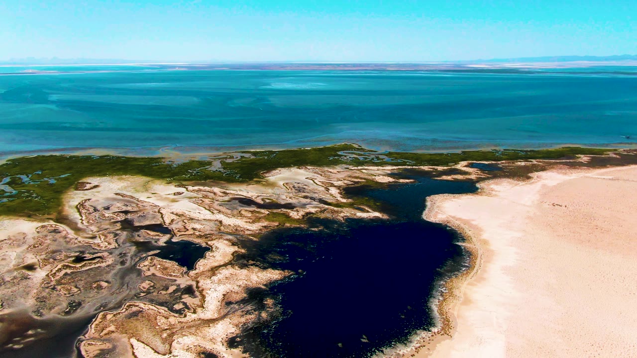 Aerial view in Guerrero Negro showing the lagoon centered lower in frame, mottled rocky flats to the left, a broad sandy shore to the right, ocean bands beyond, and no visible people.