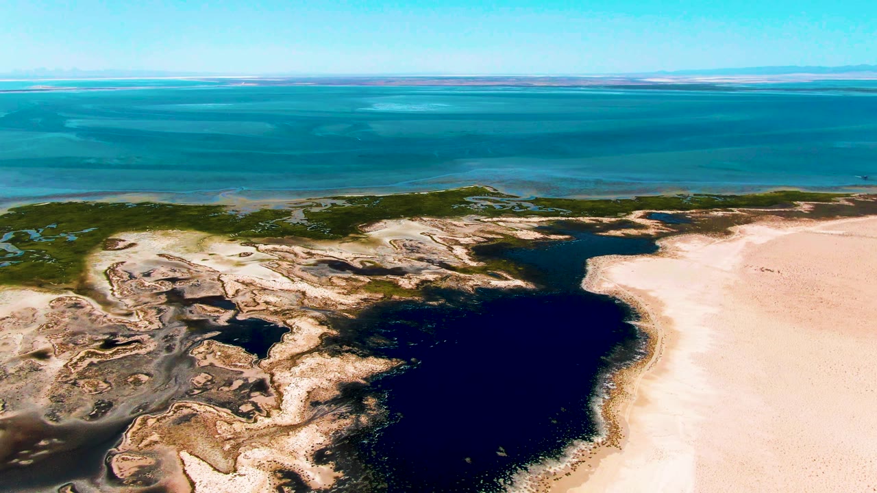 Aerial Guerrero Negro frame with the dark lagoon slightly right of center, pale sand sweeping up the right side, low green vegetation near the shoreline, turquoise ocean, and no visible people.