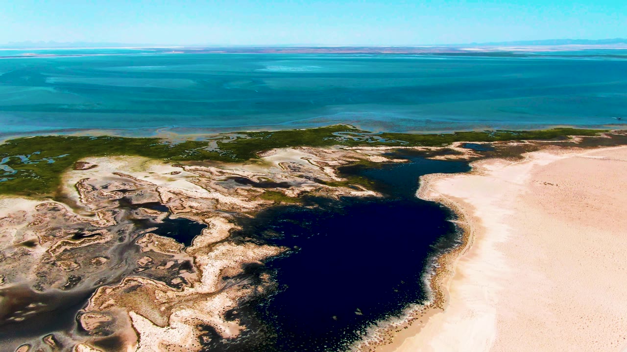 Mid-clip aerial frame over Guerrero Negro with dark lagoon water filling the lower right, textured rock and wet sand at left, pale beach at right, blue-green ocean beyond, and no visible people.