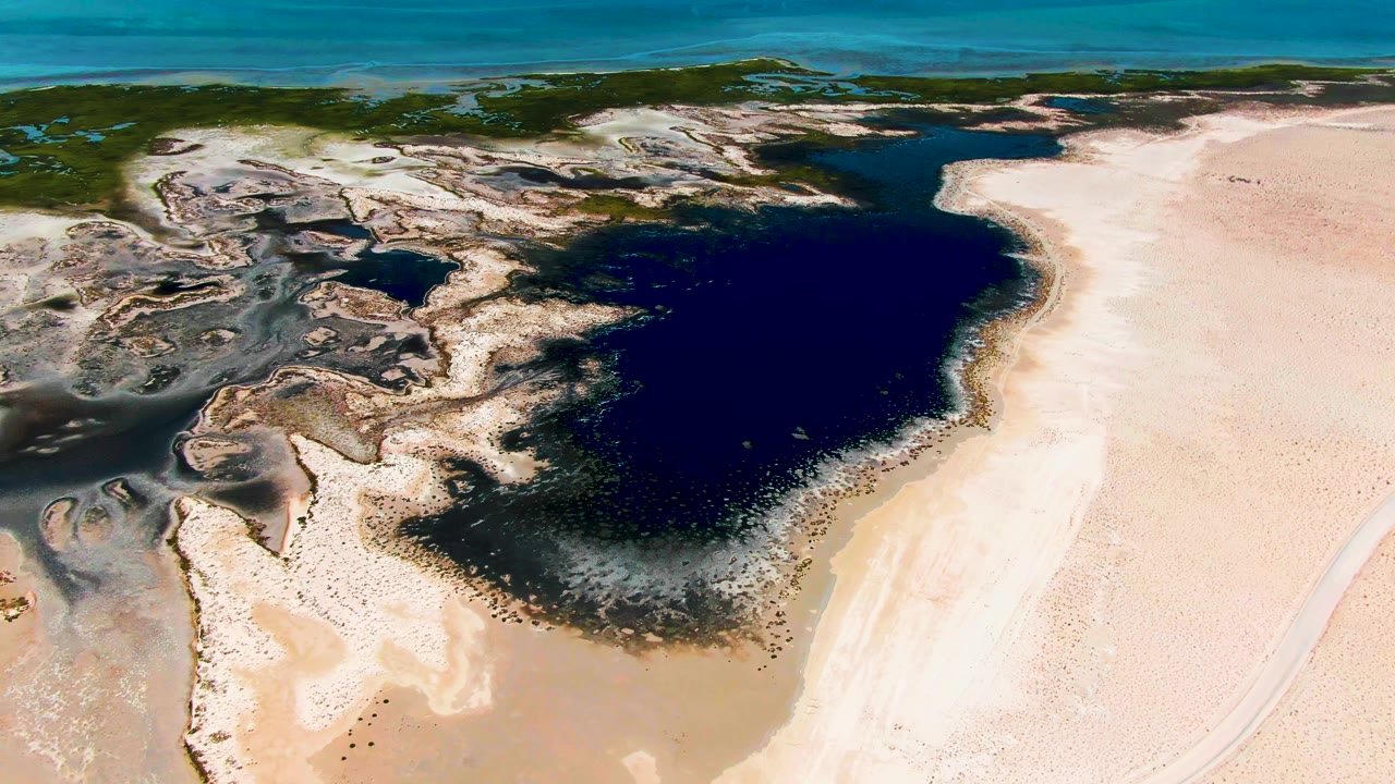 Aerial frame over Guerrero Negro with the lagoon centered, textured rocky shallows to the left, wide pale sand to the right, and no visible people.