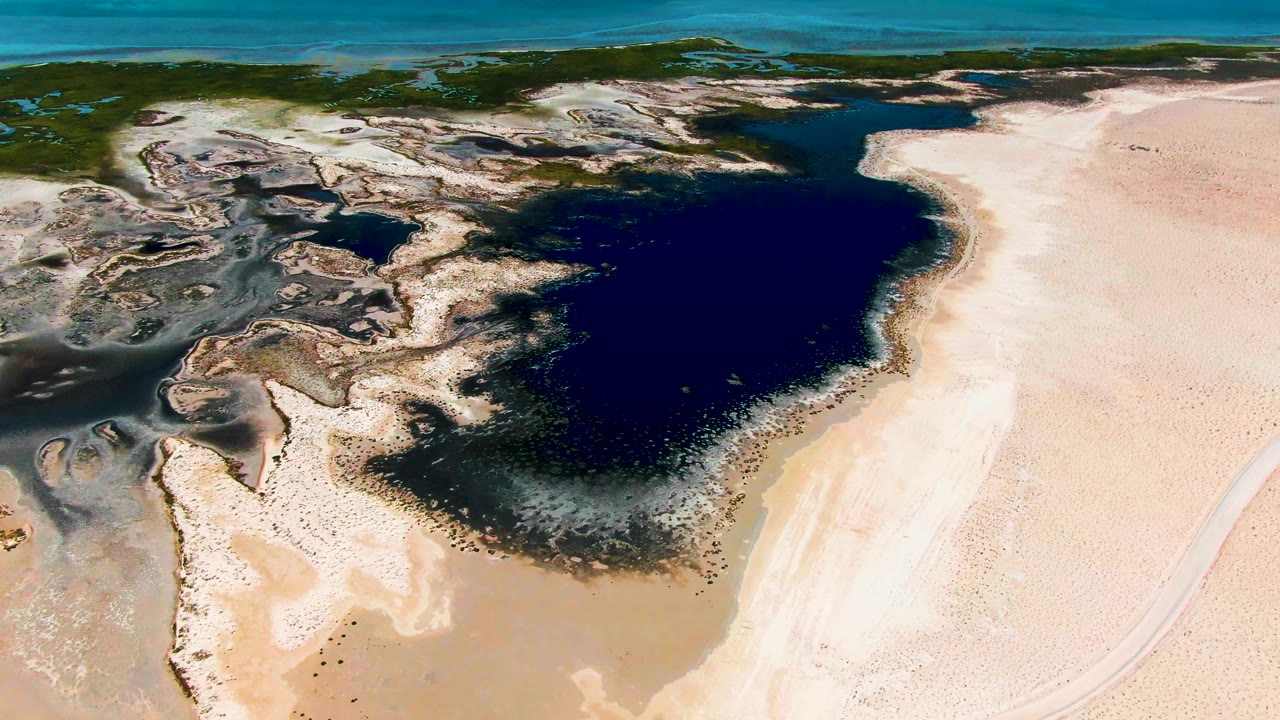Aerial frame over Guerrero Negro showing a broad dark lagoon pocket, pale sand flats on the right, mottled mudflats on the left, ocean beyond, and no visible people.