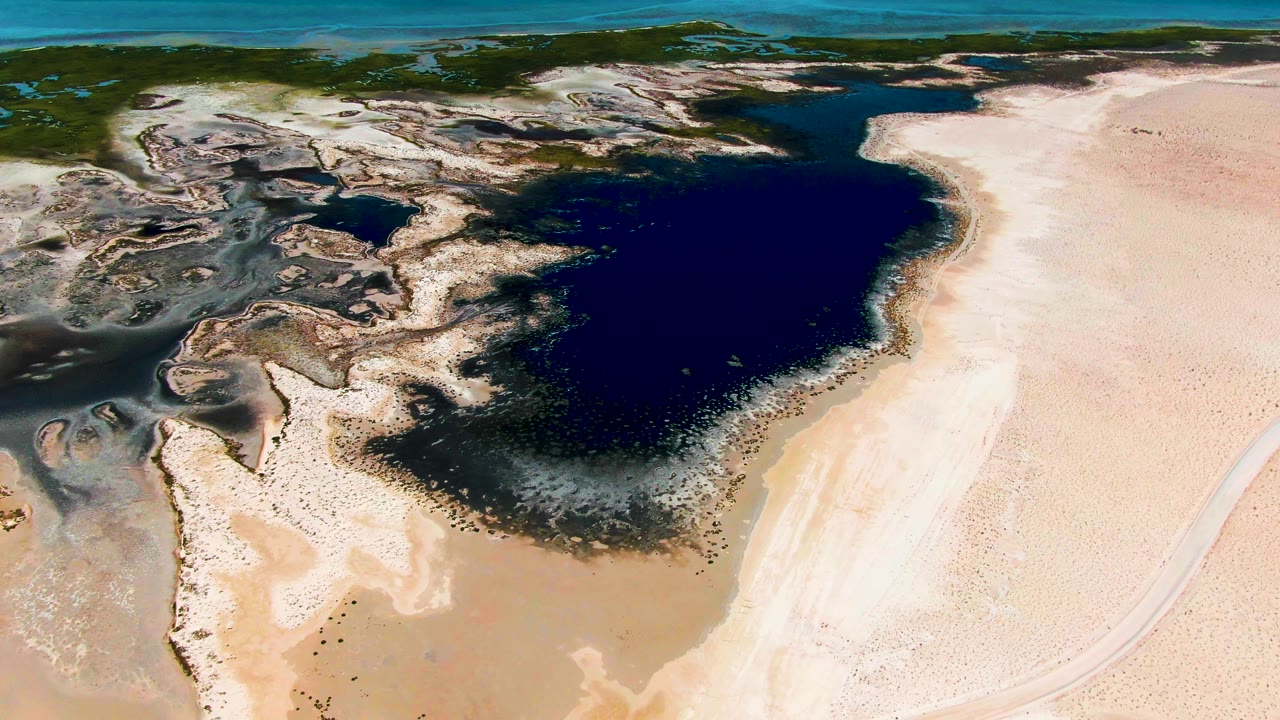 Aerial frame over Guerrero Negro with the lagoon shifted slightly right in the composition, pale sand across the lower and right edges, rocky wetland texture to the left, and no visible people.