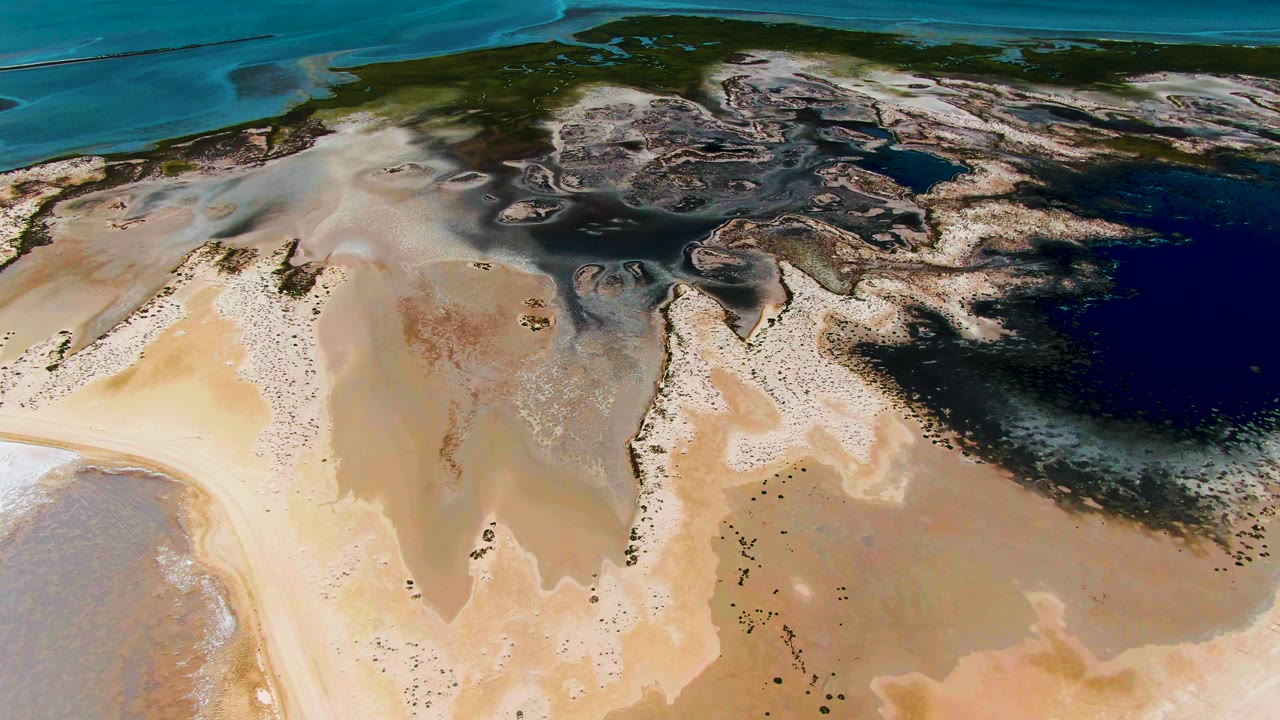 Aerial view over Guerrero Negro tidal flats with a central tan sand area, dark shallow pools to the right, turquoise water in the background, and no visible people.