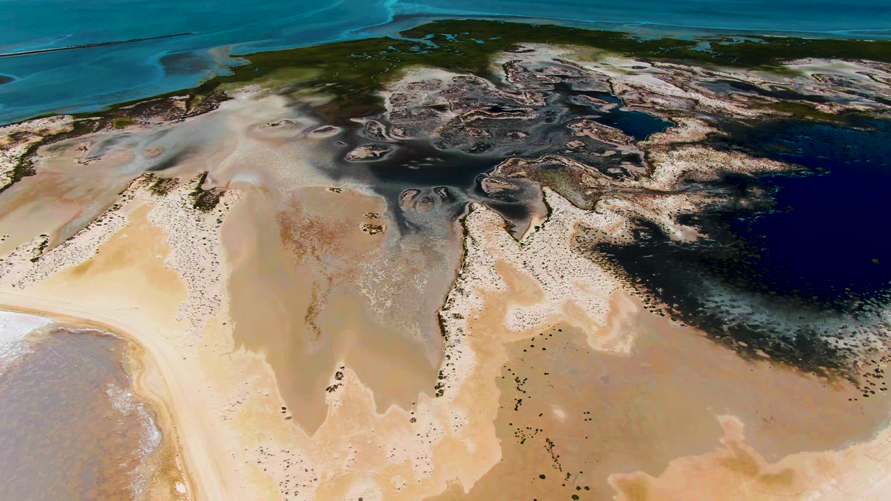 High aerial frame in Guerrero Negro showing pale sand flats stretching toward blue-green coastal water, with darker wet areas and no visible people.
