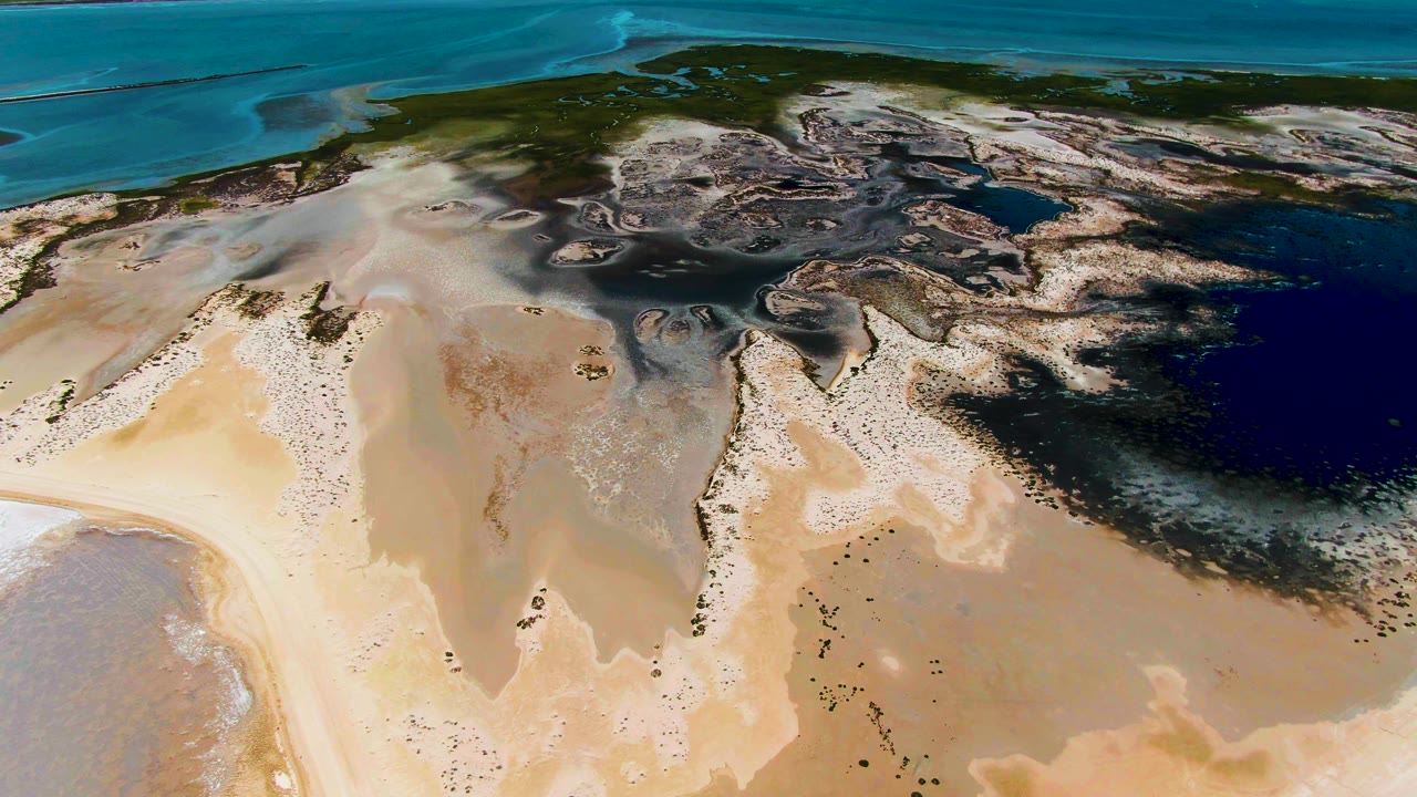 Aerial frame from Guerrero Negro with a wide pale sandbar in the lower frame, dark tidal flats across the middle, blue water beyond, and no visible people.