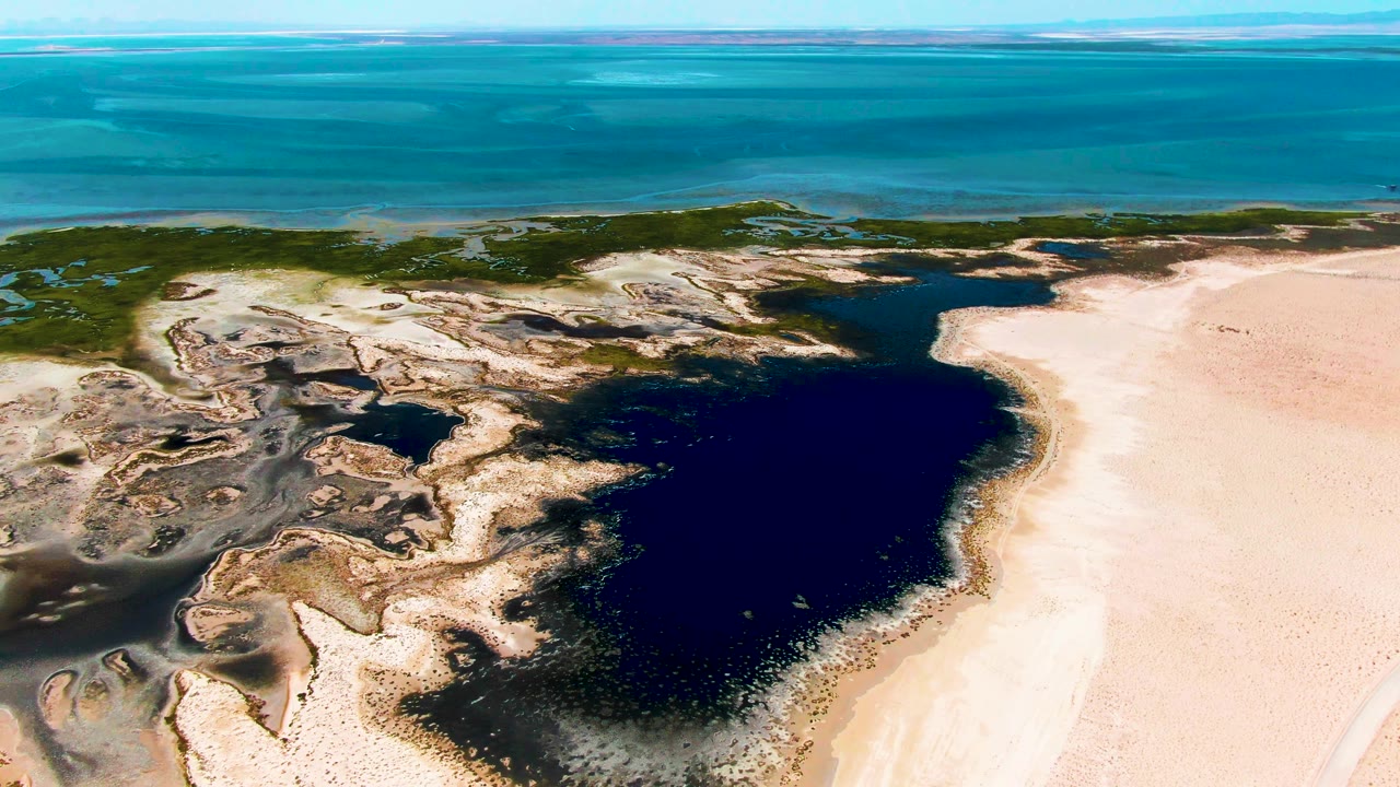 Aerial frame over Guerrero Negro with the lagoon centered more prominently, pale sand to the right, textured mud flats to the left, and turquoise ocean across the background, no visible people.