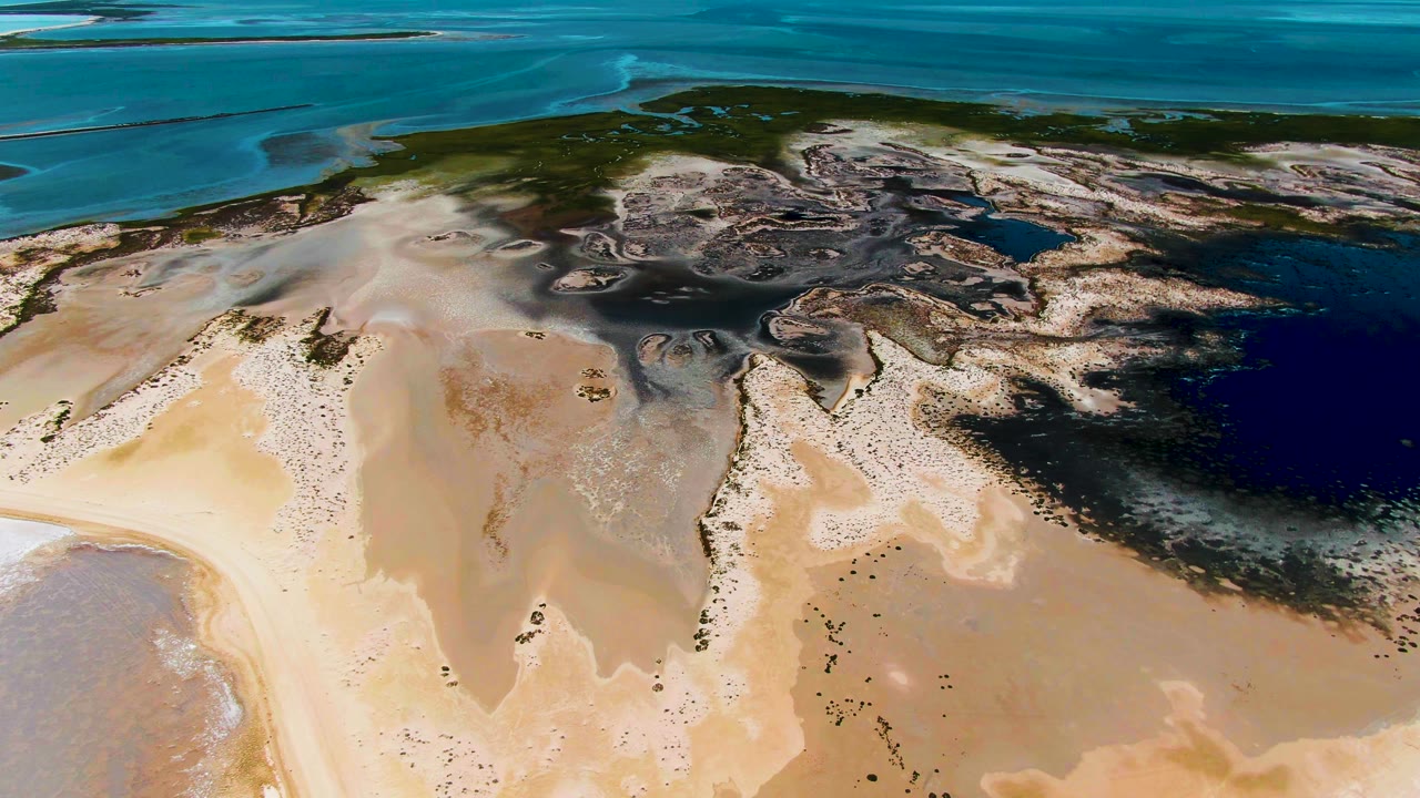 Opening aerial frame over Guerrero Negro, Baja California Sur, showing pale sand tidal flats below dark wetland bands and blue coastal water beyond, with no visible people.