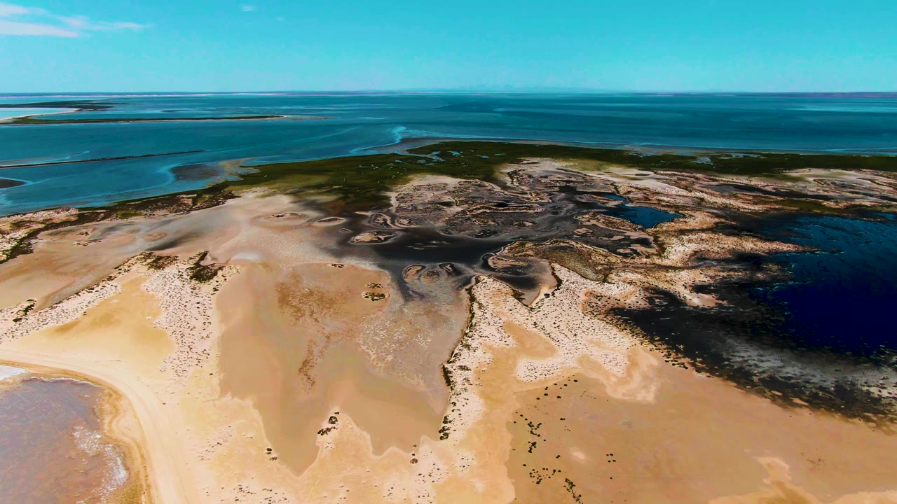 Aerial coastal landscape in Guerrero Negro with pale sand in the lower frame, scattered tidal pools and wet marks, a green midground strip, blue sea beyond, and no visible people.