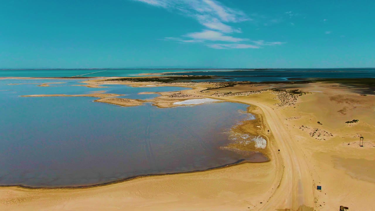Aerial frame over Guerrero Negro, Baja California Sur, showing tan shoreline, shallow tidal pools, a sandy dirt road, distant ocean, scattered clouds, and no visible people.
