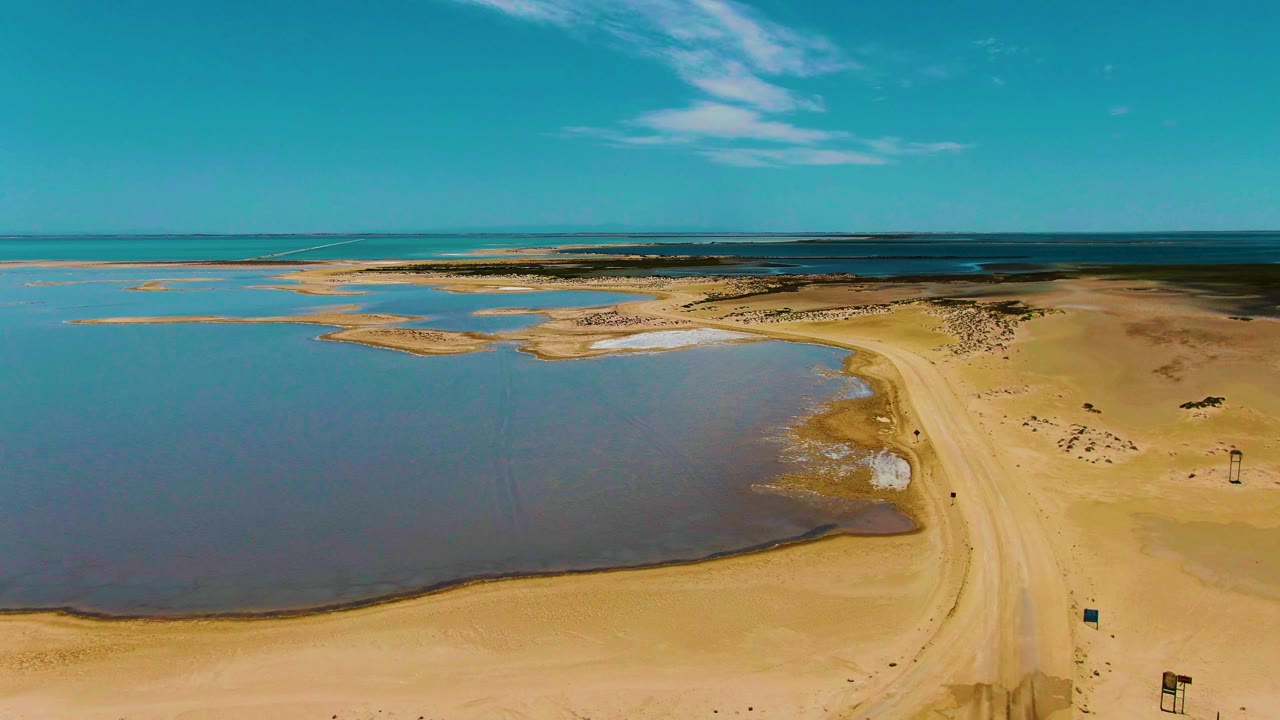 Second aerial frame in Guerrero Negro with the shallow pool filling the left side, the beach road curving through the right side, the ocean on the horizon, and no visible people.
