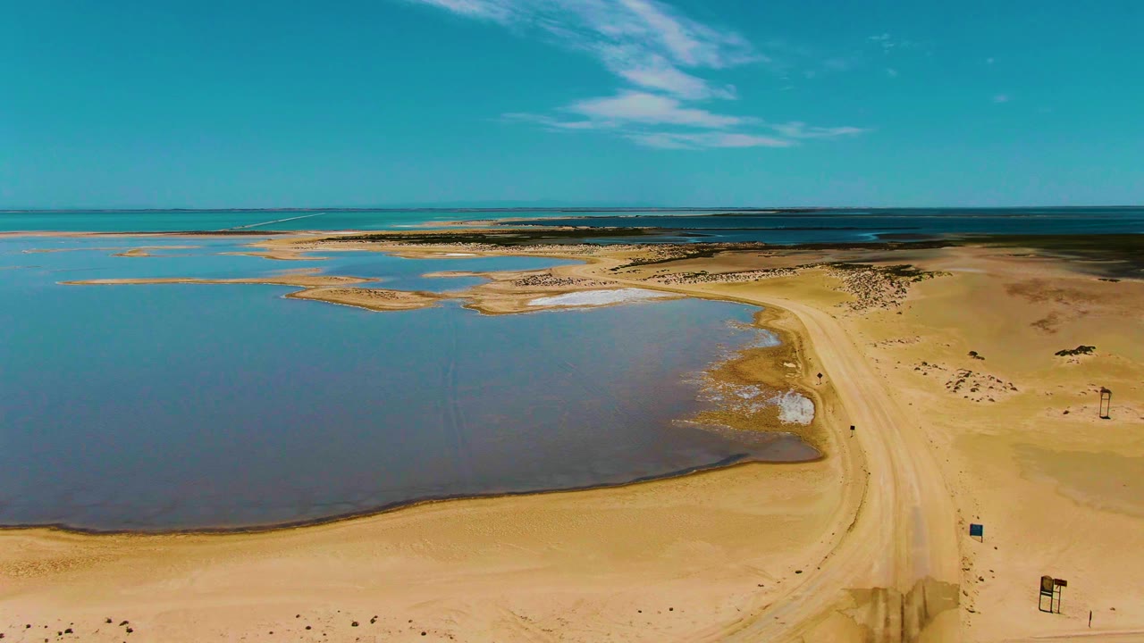 Aerial view over Guerrero Negro with blue shallow water along the left foreground, pale sand bars near the center, a dirt track on the right, and no visible people.