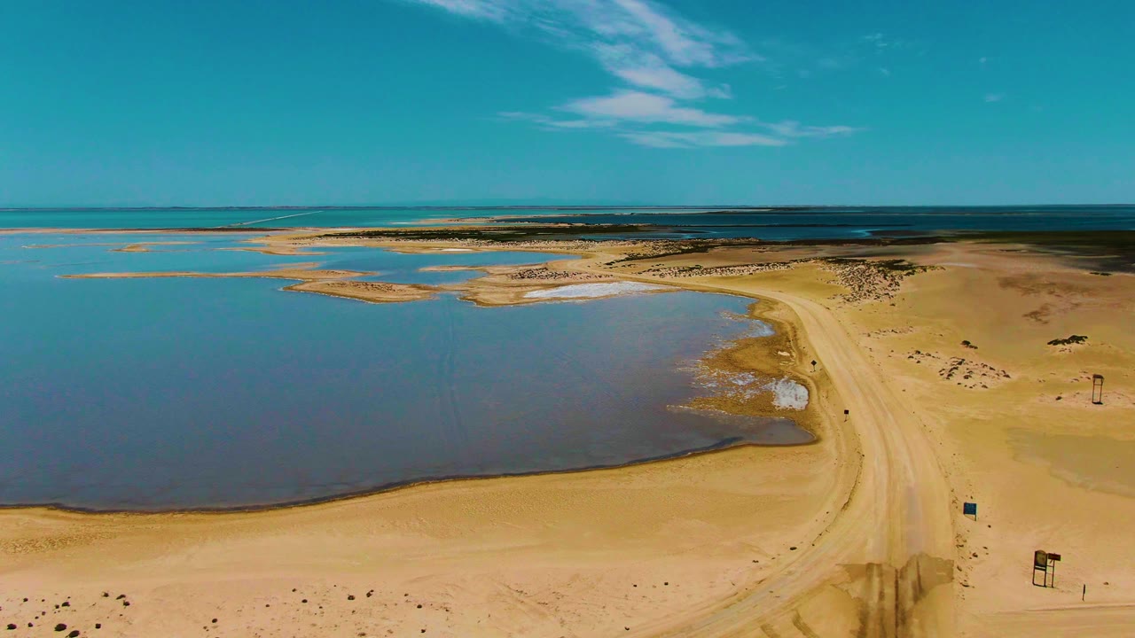 Coastal desert shoreline with tidal pool, sandy road, and open sky.