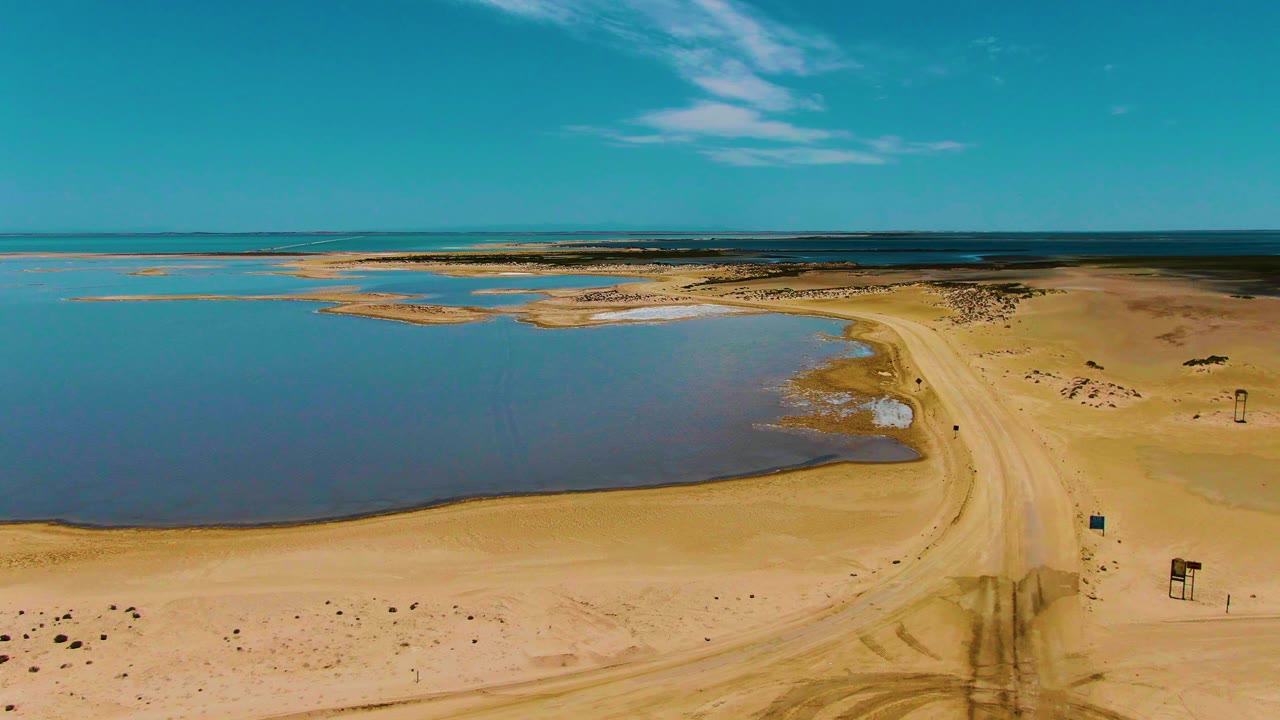 Aerial view of a sandy road curving beside blue water under a clear sky.