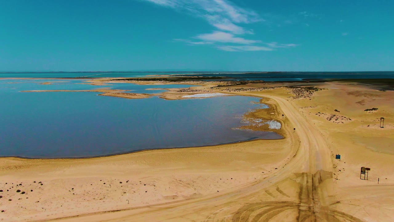 Aerial view in Guerrero Negro, Baja California Sur, with dirt tracks crossing pale sand toward a curved road beside calm blue water and no visible people.